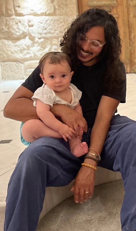 Beez sitting on the floor holding his baby son on his lap, smiling at the camera